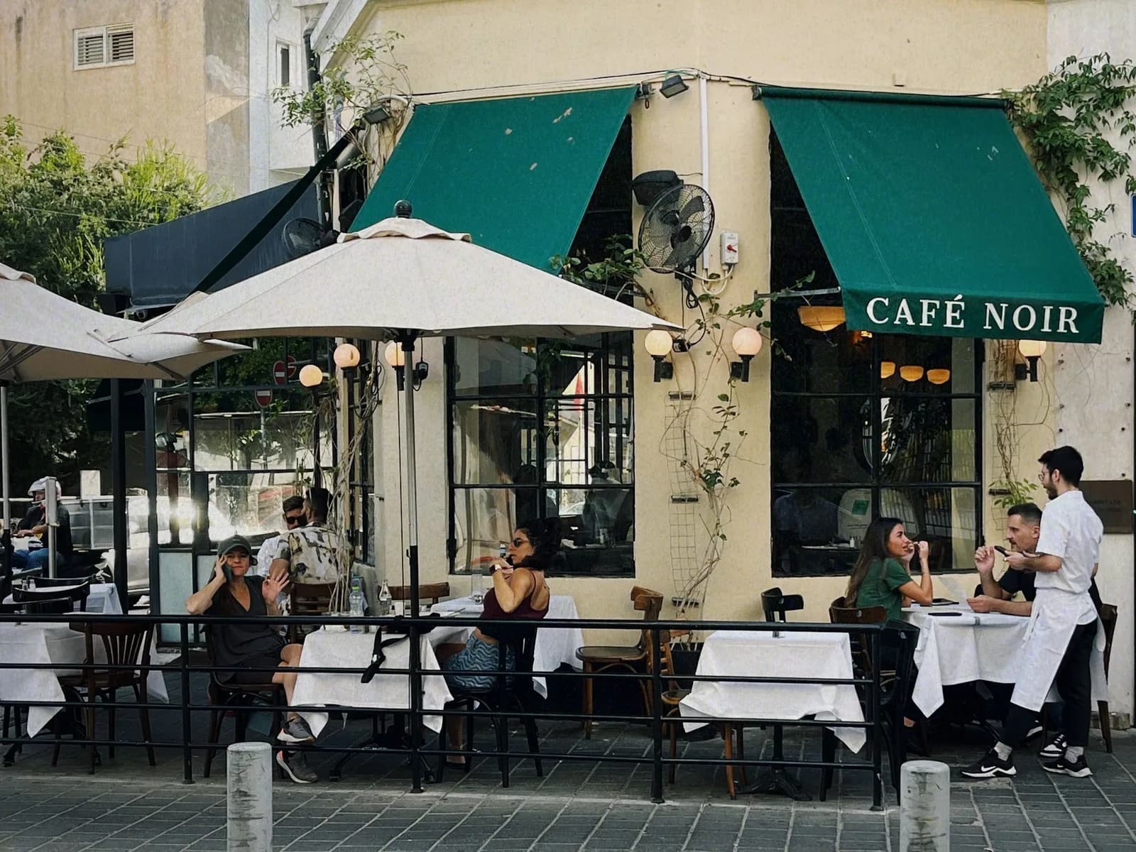 A street café in Tel Aviv in daylight, tables under green awnings and customers seated outside