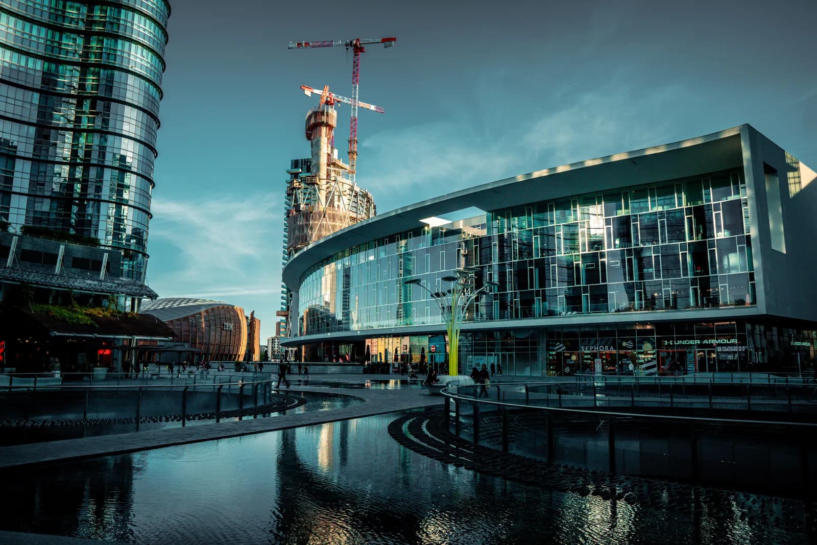 The Porta Nuova business district in Milan: glass office towers, a curved retail building, a construction crane and people walking beside the water